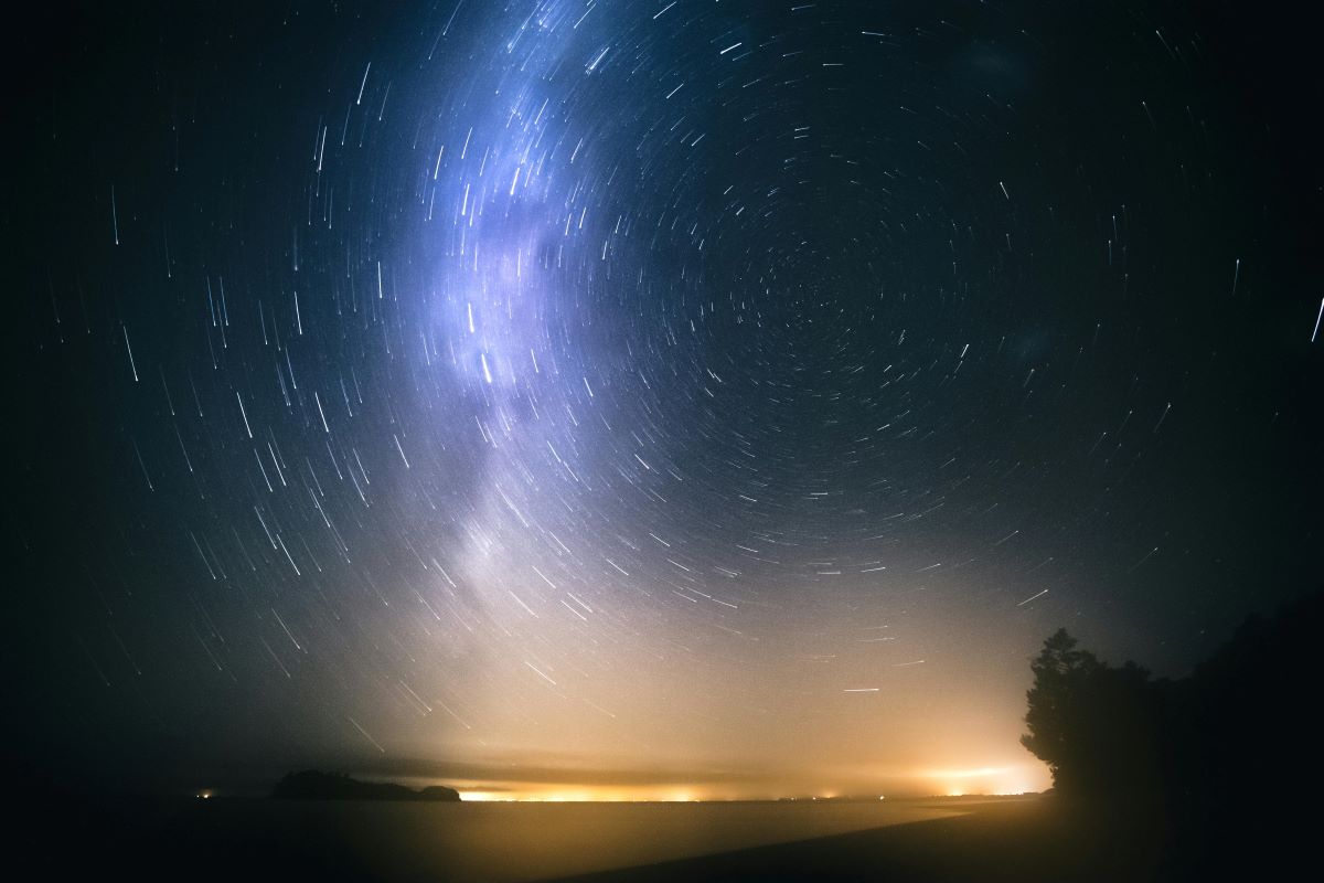 A long-exposure night sky photograph shows circular star trails above a coastal landscape illuminated by distant city lights.