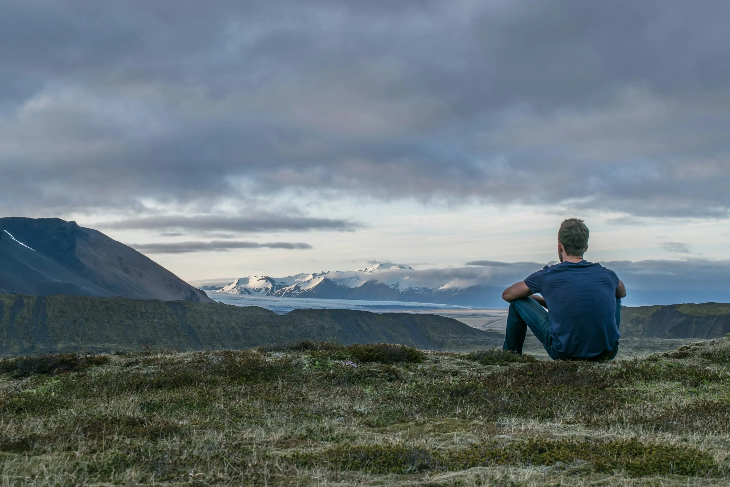 Person sitting on grassy hill overlooking mountains and cloudy sky, reflecting in a calm, contemplative moment